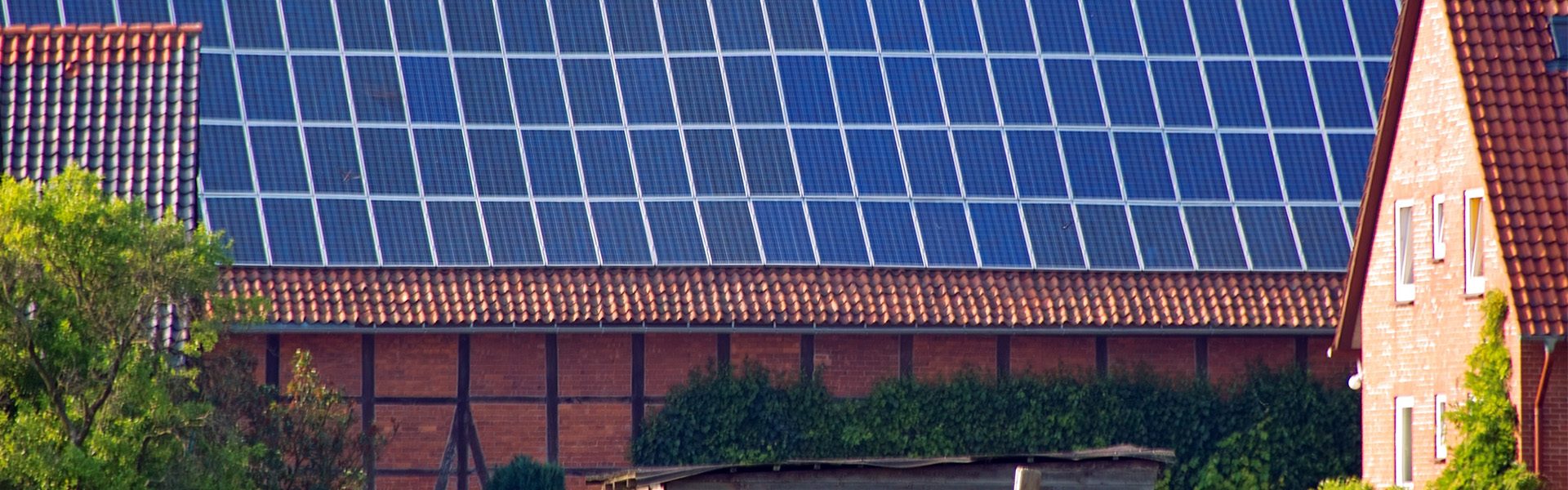 Das Bild zeigt ein Gebäude mit einer großen Fläche an Solarpaneelen (Photovoltaikanlage) auf dem Dach. Rechts und links im Bild sind außerdem Teile von roten Hausdächern mit Dachziegeln zu erkennen.