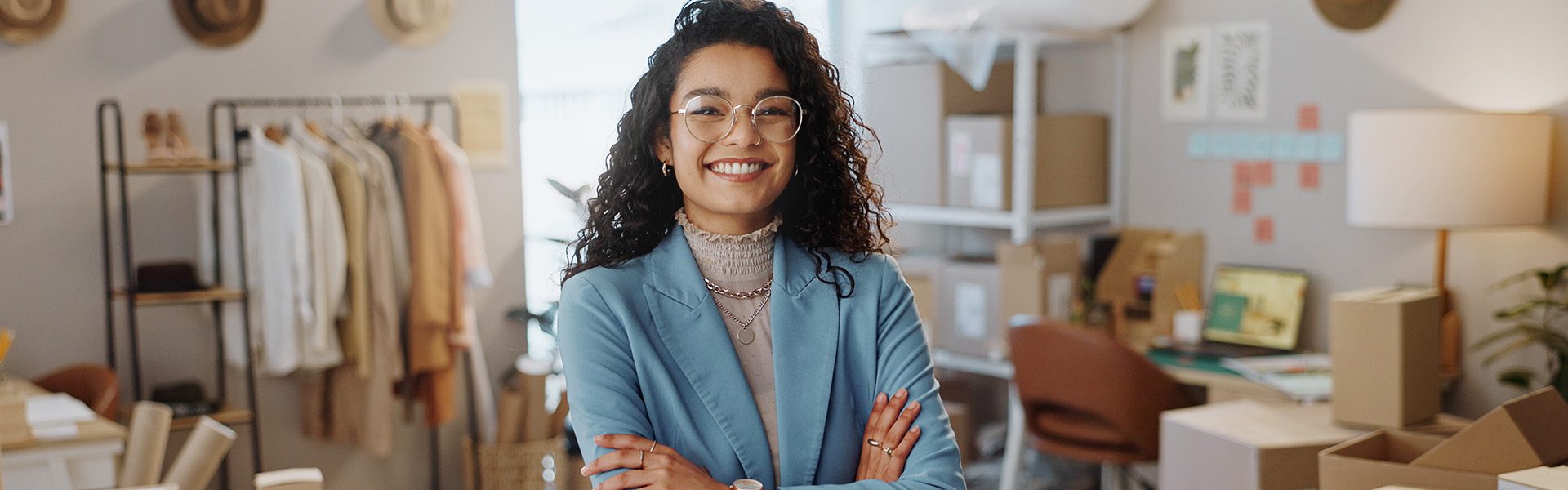 Das Bild zeigt eine Person in einem Raum, der wie ein Büro oder Arbeitsplatz aussieht. Im Hintergrund sind Kleider auf einem Ständer, Kartons und Büromöbel zu sehen. Es erweckt den Eindruck eines kreativen Arbeitsplatzes oder eines Online-Geschäftsumfelds.