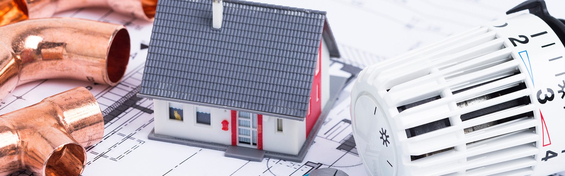 Das Bild zeigt ein kleines Modell eines Hauses auf technischen Zeichnungen, zusammen mit Heizungsrohren und einem Heizkörperthermostat. Es symbolisiert Themen rund um Heizungstechnik, Hausbau oder Haustechnik.