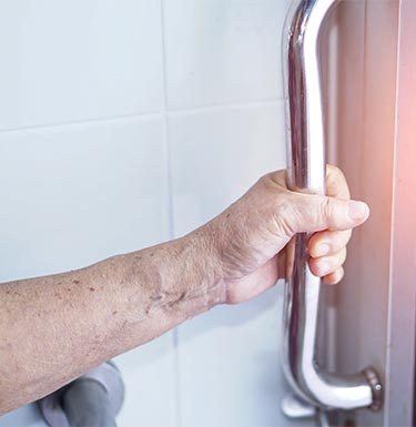 Das Bild zeigt eine ältere Person, die einen stabilen Griff oder Haltegriff in einer Dusch- oder Badezimmerumgebung hält. Dieser Griff ist in der Regel aus Metall und wird häufig in Badezimmern installiert, um zusätzliche Sicherheit und Unterstützung zu bieten, besonders für ältere Menschen oder Personen mit eingeschränkter Mobilität. Der Griff dient dazu, beim Ein- und Aussteigen aus der Dusche oder der Badewanne sicheren Halt zu gewährleisten, um mögliche Ausrutscher oder Stürze zu vermeiden.
