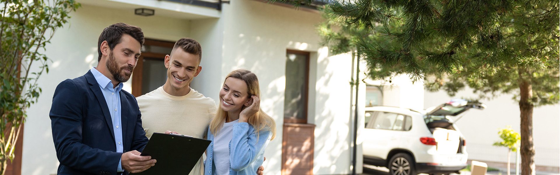 Das Bild zeigt eine Immobilienbesichtigung oder eine Beratung vor einem Haus. Eine Person in einem Anzug hält eine Mappe, während zwei andere Personen daneben stehen und ihm zuhören. Im Hintergrund ist ein geparktes Auto sichtbar.