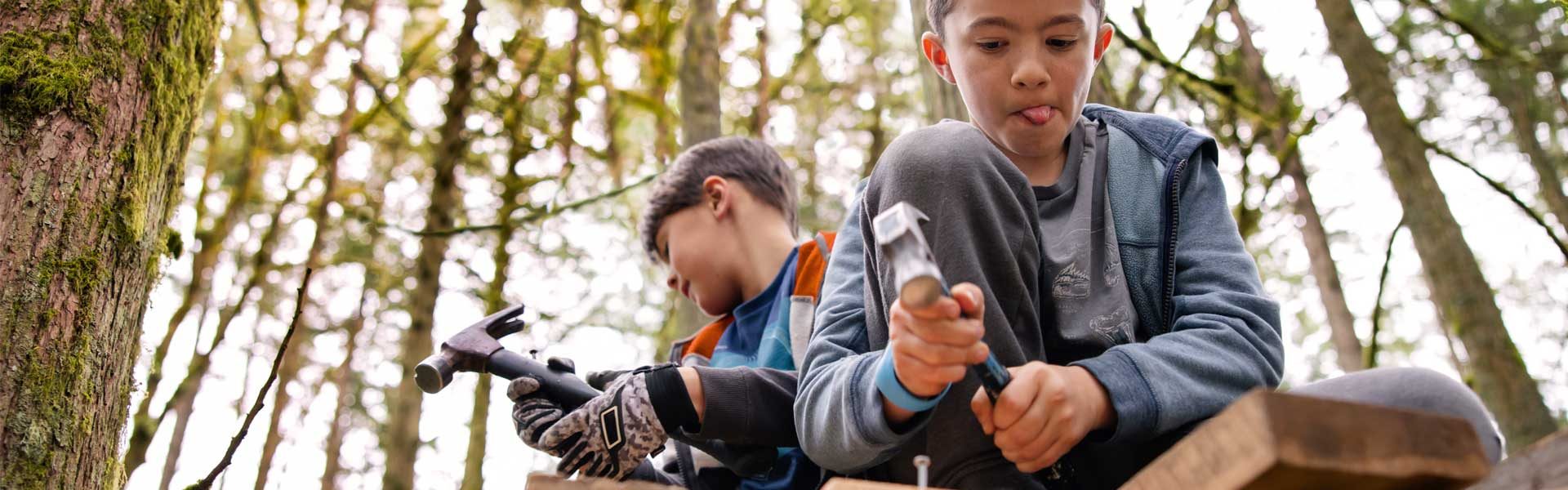 Das Bild zeigt zwei Kinder, die auf einer Holzplattform oder einem Holztisch im Freien sitzen, umgeben von Bäumen und einem bewaldeten Gebiet. Sie sind in Freizeitkleidung und scheinen in eine Tätigkeit oder ein Spiel vertieft zu sein.